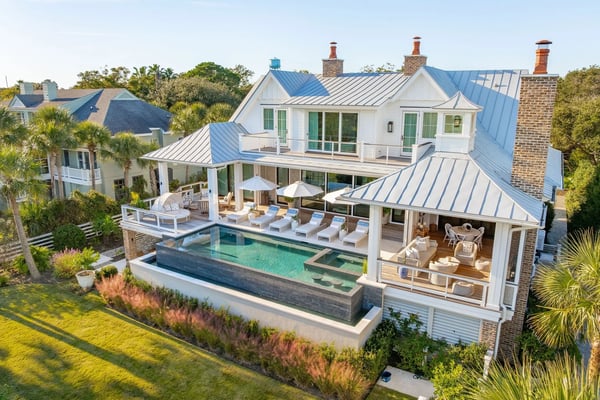 Modern white farmhouse with blue metal roof, pool, lounge chairs, and brick chimney surrounded by palm trees and manicured lawn.